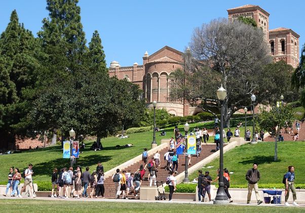 Students walk across UCLA's sun-drenched campus with Royce Hall's distinctive red brick towers visible in the background.