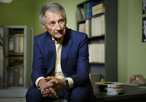 Leonard Kleinrock, wearing a navy suit and pale yellow shirt, sits with his hands clasped in an office, with bookshelves, computer equipment and a vintage telephone behind him.