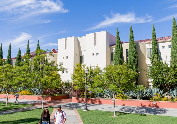 Two people walk along a tree-lined pathway beside a UCLA residential building surrounded by landscaped greenery.