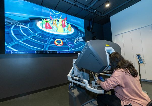Seated woman bends her head into surgery simulation machine. A video screen with surgical graphics is in the background.