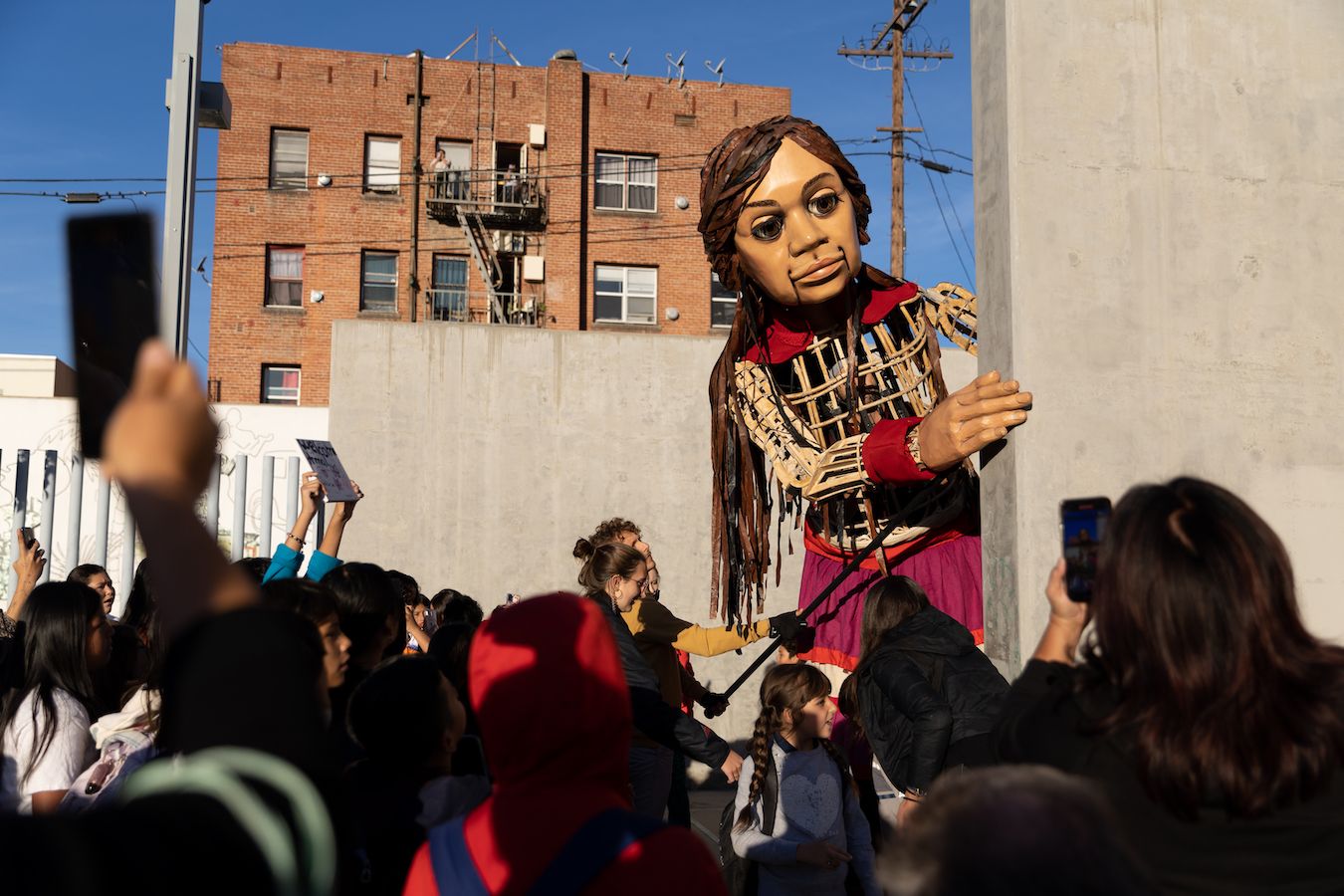Giant puppet ‘Little Amal’ peaks around the corner of a building as students watch on