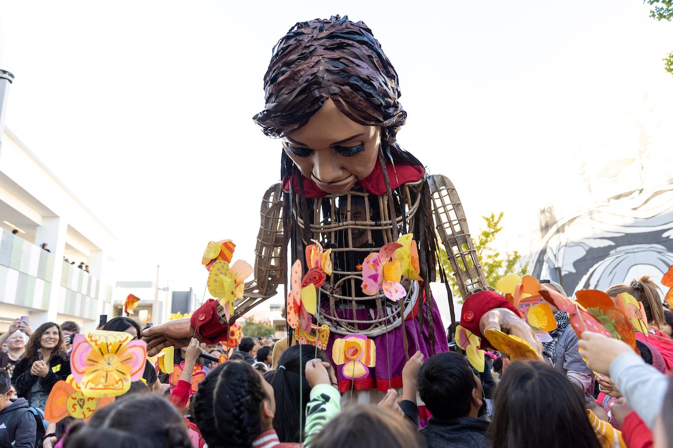 Giant puppet ‘Little Amal’ bends down toward a large group of students holding paper poppies