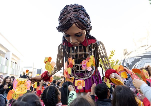 Giant puppet ‘Little Amal’ bends down toward a large group of students holding paper poppies