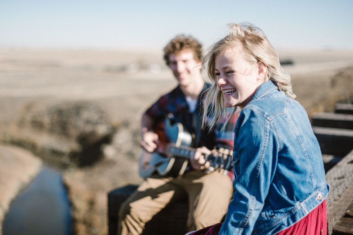 Person wearing denim jacket smiling as another person in plaid shirt strums guitar