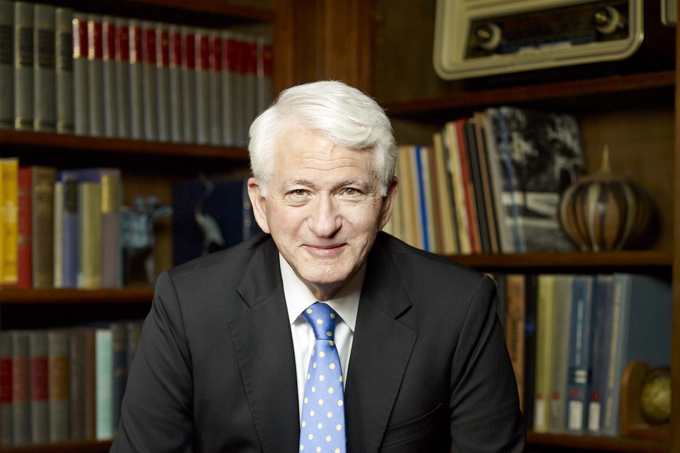 Portrait of Gene Block in black blazer and blue polka-dot tie with bookcase in background