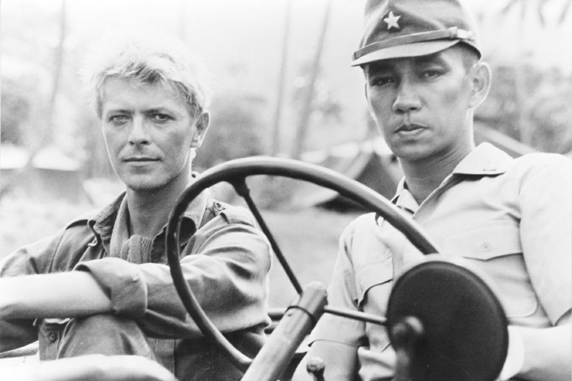 Black-and-white photo still from “Merry Christmas, Mr. Lawrence” featuring David Bowie and Ryuichi Sakamoto in an army jeep