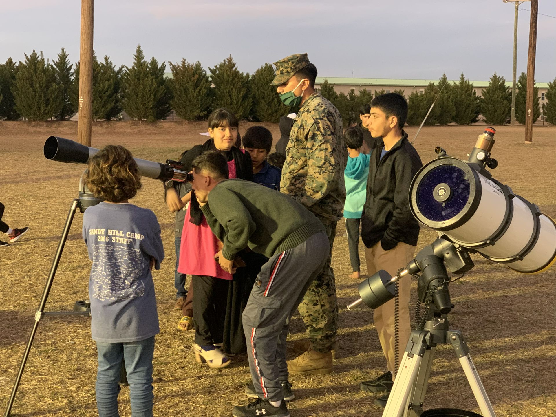Several young children stand around man in military fatigues and look through telescopes