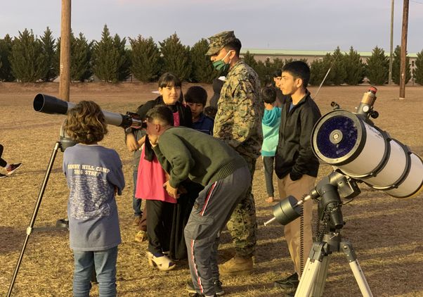 Several young children stand around man in military fatigues and look through telescopes
