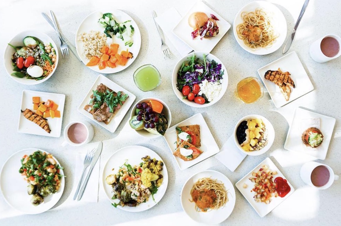 A small sampling of the fresh, colorful, healthy meal selections offered at UCLA. View from above of about a dozen dishes on a white table top.