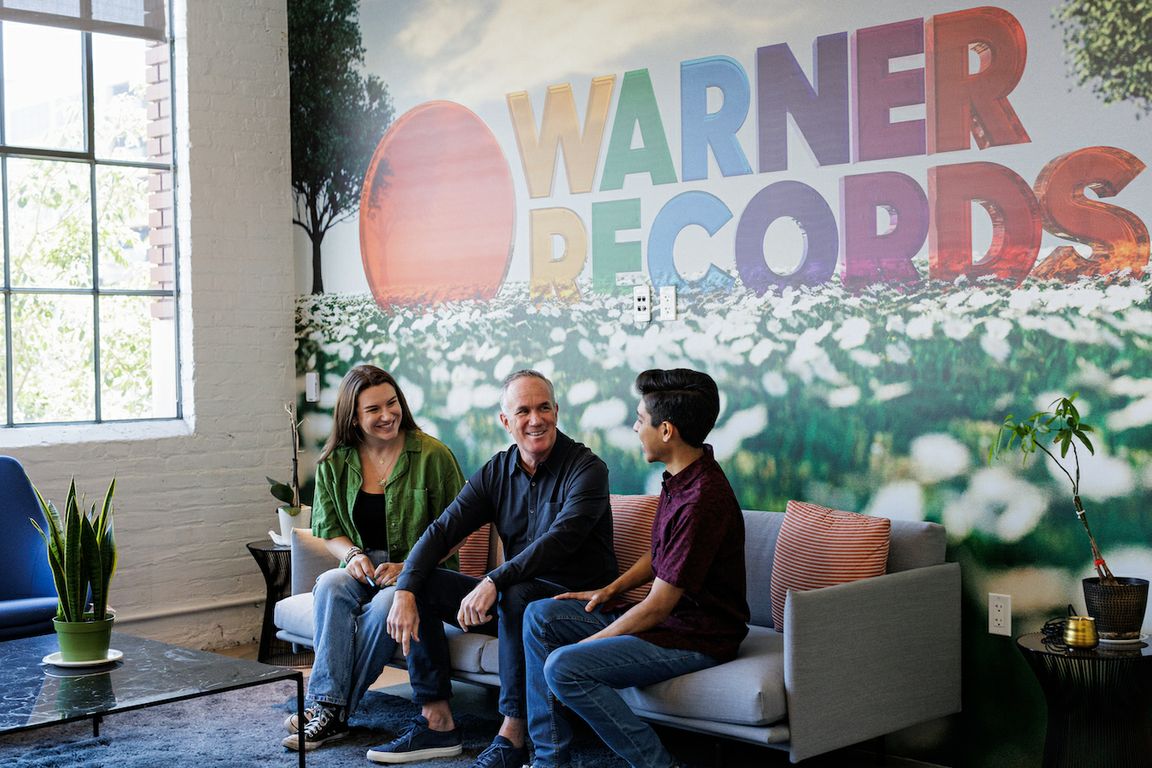 Three people seated on couch beneath Warner Records sign