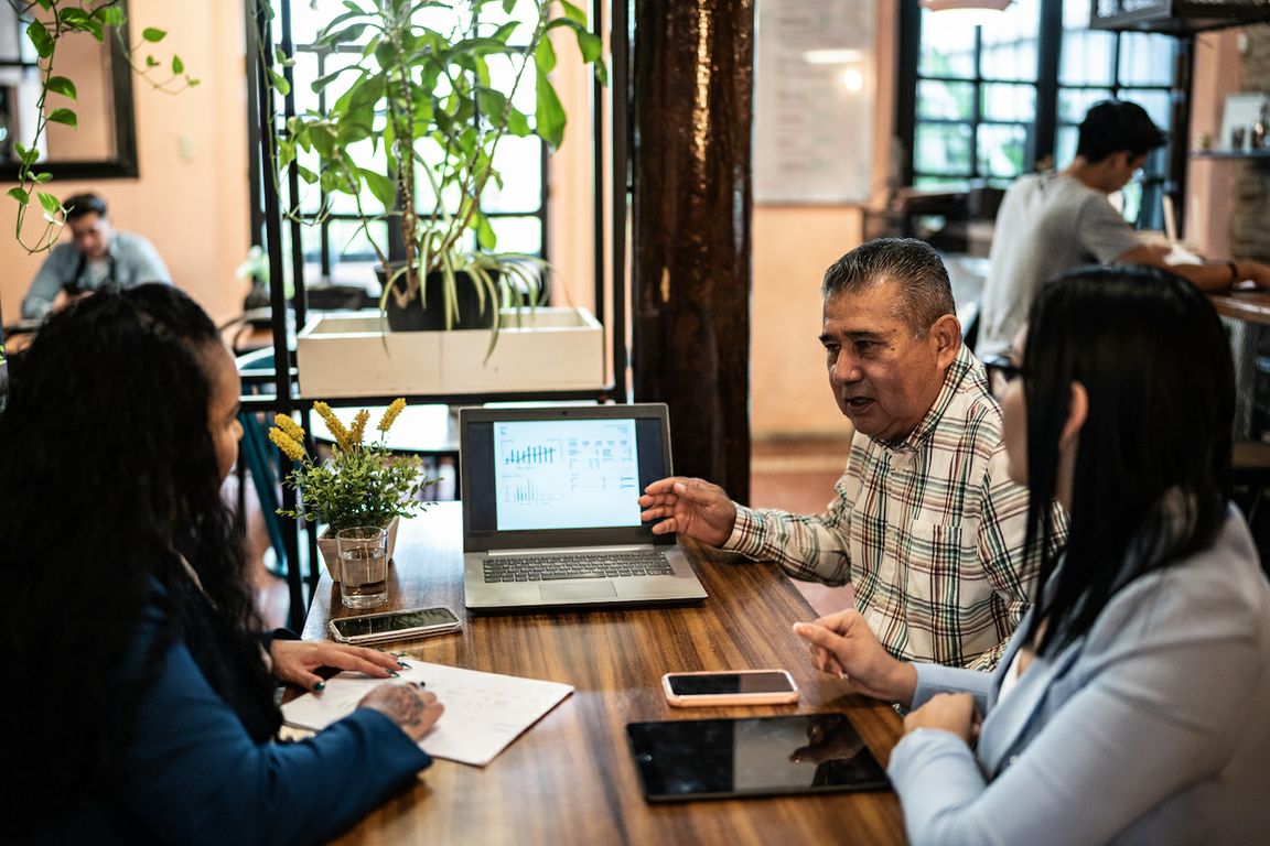 Three Latinos of varying ages sitting at a table with a laptop, pad of paper, phone and tablet