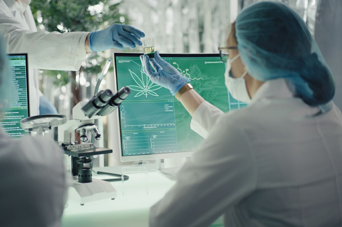 Scientist in lab, in front of miscroscope and computer screen featuring cannabis leaf, takes a vial from another scientist’s hand