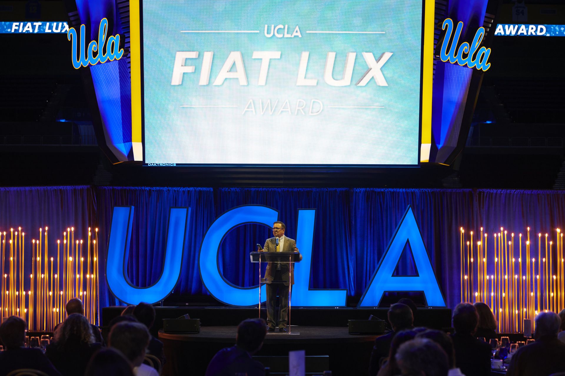 Wide shot of Tony Pritzker speaking at podium on stage at Pauley Pavilion. Behind him are a video screen reading “UCLA Fiat Lux Award” and large block letters spelling out “UCLA.” 