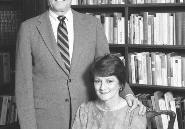 Black-and-white image of Chancellor Block standing next to his wife, Sue, who is seated a desk. In the background is a bookcase