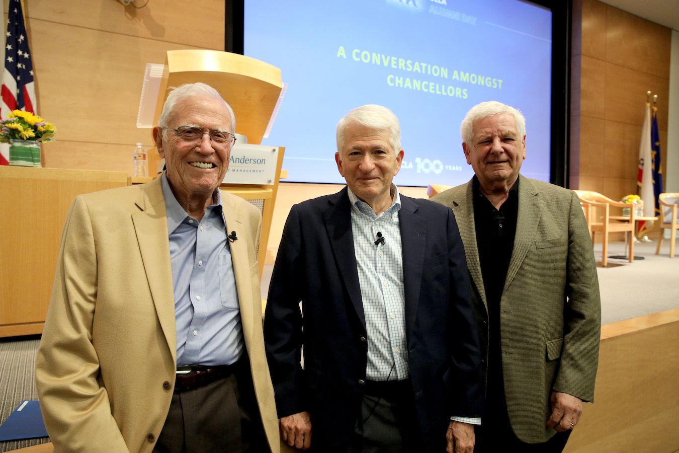 Left to right: Charles E Young, Gene Block and Albert Carnesale stand in front of a stage, with a screen in the background reading, “A Conversation Amongst Chancellors.”