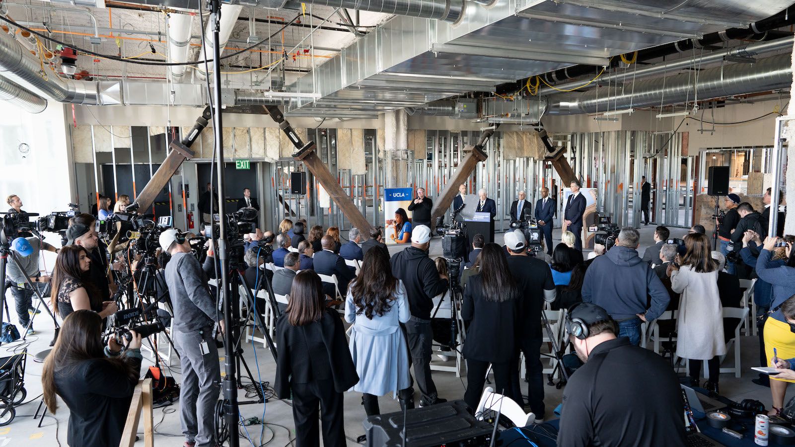 Wide shot with members of press and supporters. Chancellor Block speaks at podium in the background