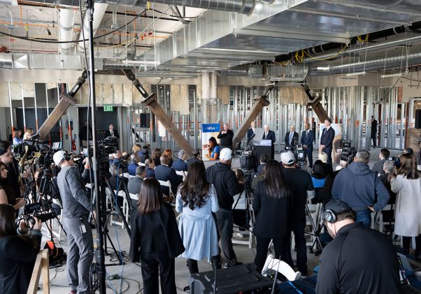 Wide shot with members of press and supporters. Chancellor Block speaks at podium in the background