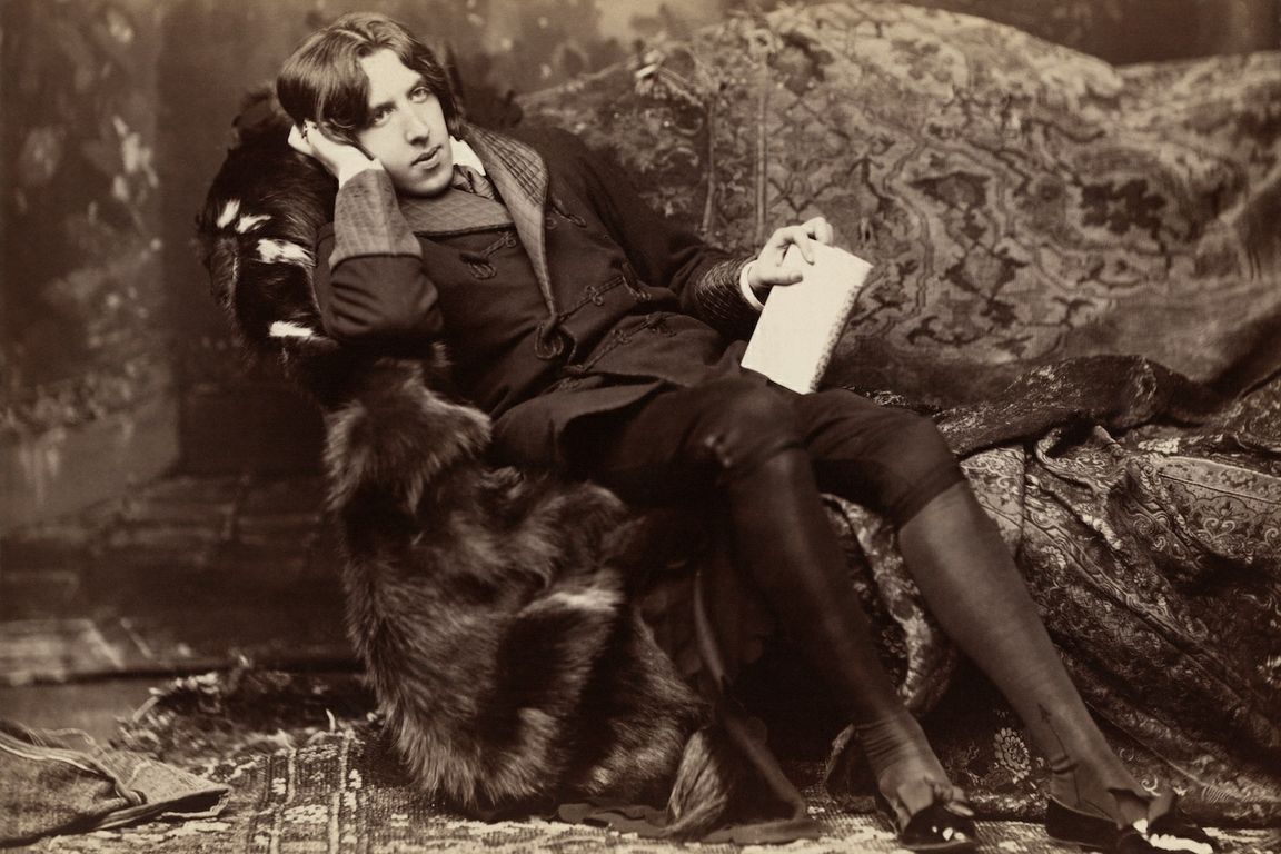 Oscar Wilde reclining on a fur-covered sofa with a book