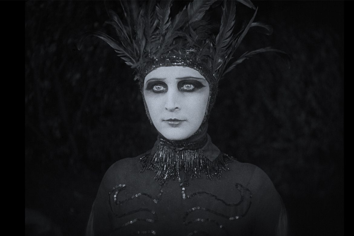 Black-and-white portrait of person wearing feathered headdress and dark eye makeup