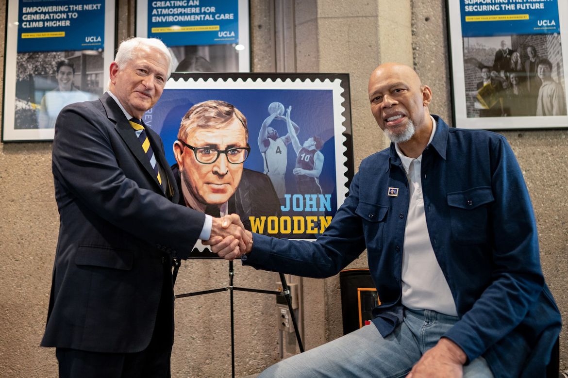 Chancellor Block and Kareem Abdul-Jabbar shake hands in front of enlarged version of Wooden stamp