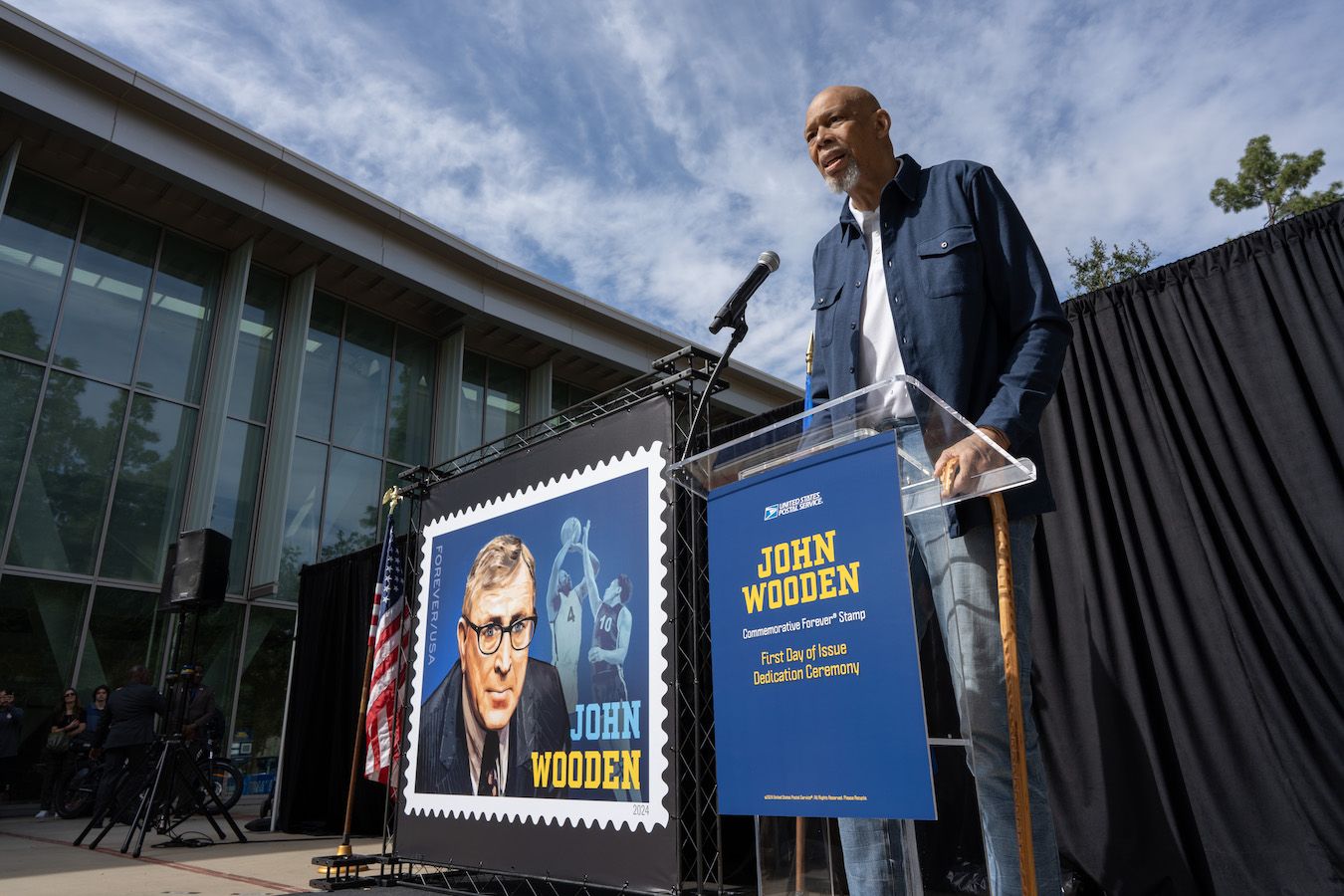 Kareem Abdul-Jabbar speaking at podium next to enlarged John Wooden stamp