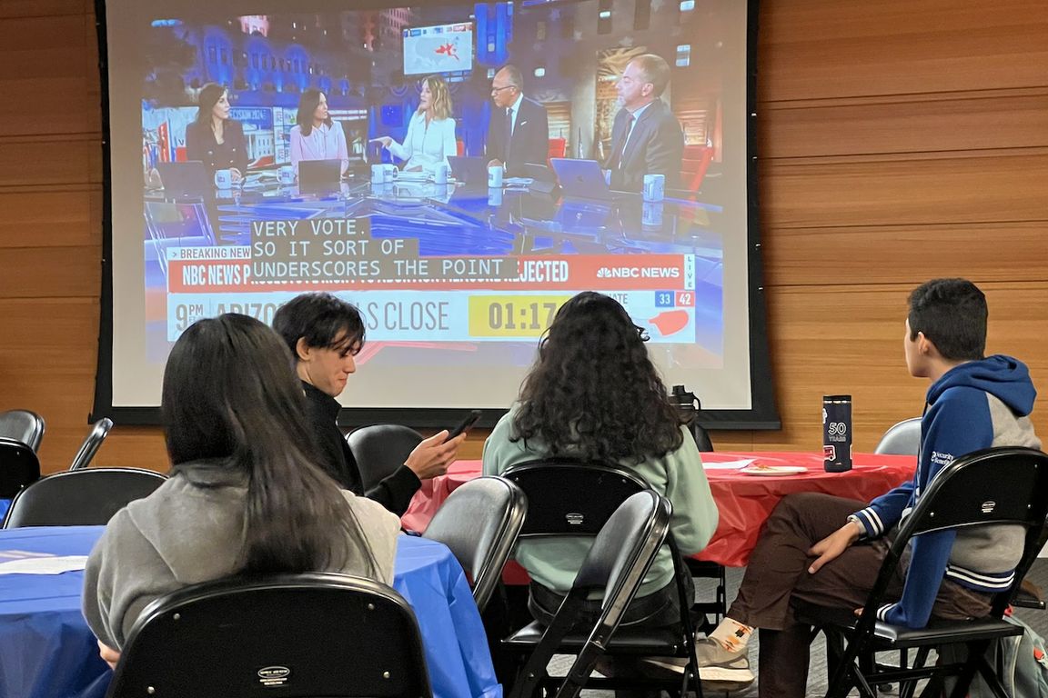 Election night watch party — group of students sits at a table in front of a TV screen displaying 2024 election results