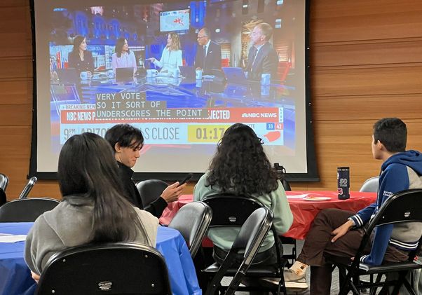 Election night watch party — group of students sits at a table in front of a TV screen displaying 2024 election results