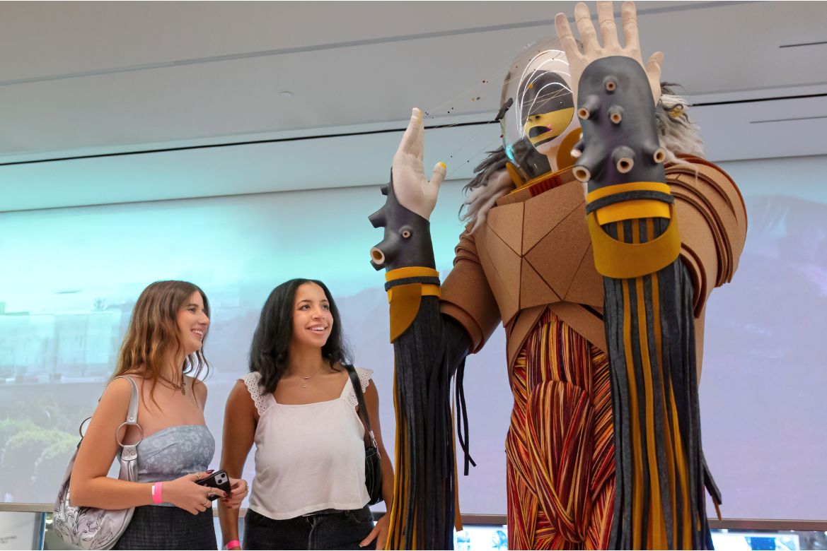 Two women react to  “Sovereign,” a large-scale Indigenous figure in a futuristic helmet and attire, standing in the Hammer Museum at UCLA lobby