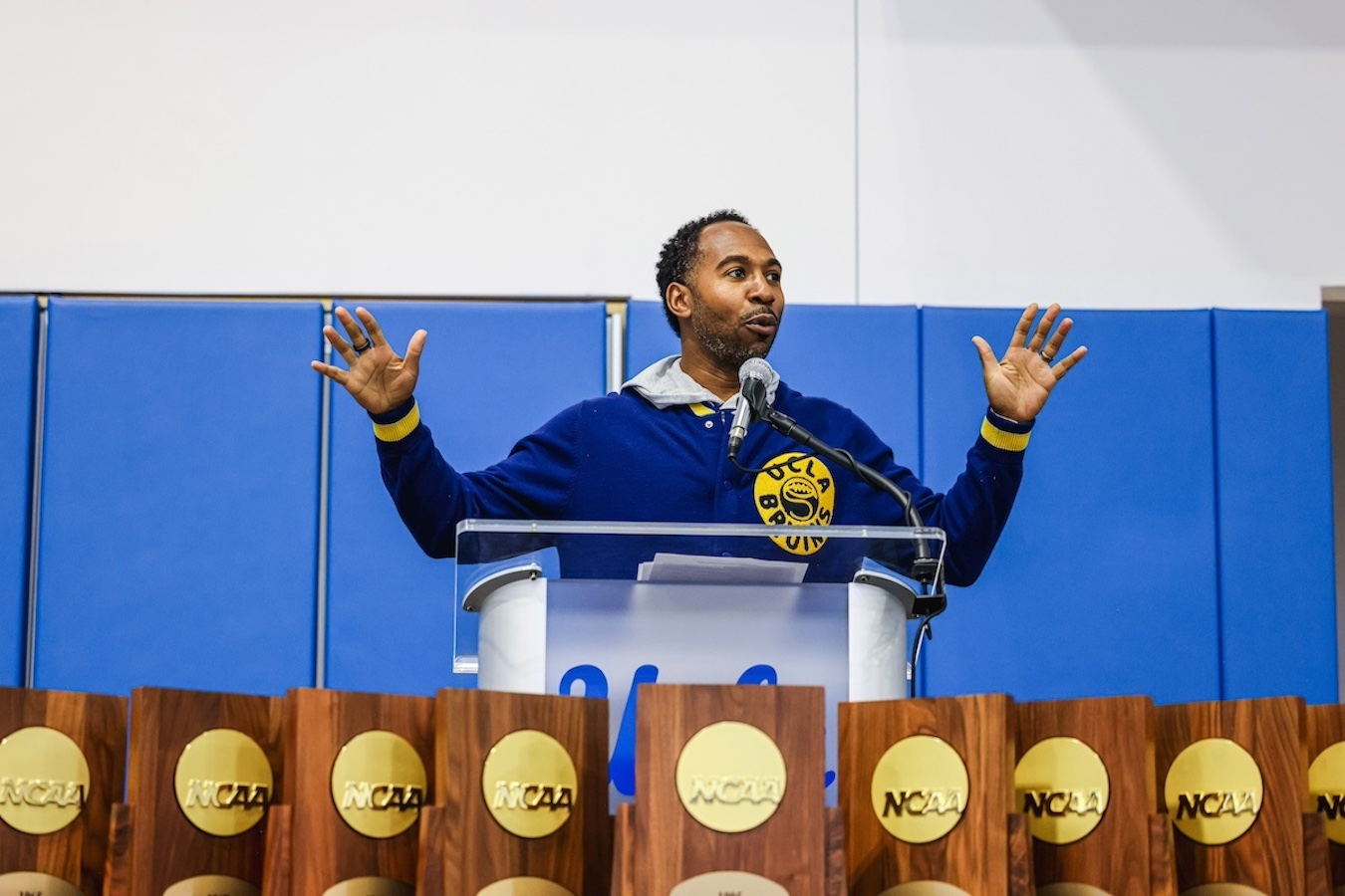 UCLA Athletics Director Martin Jarmond stands at podium in front of a line of NCAA championship trophies