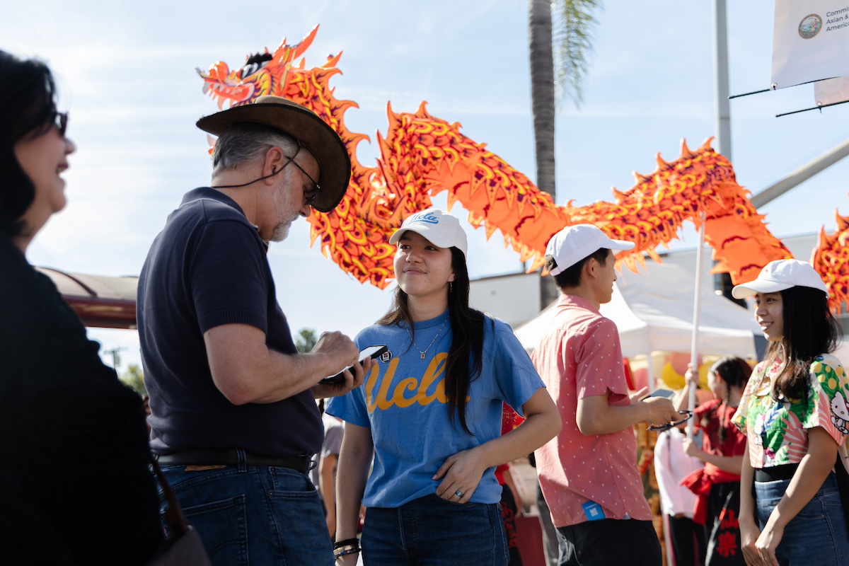 Female student in UCLA shirt administers survey to man outside, with orange paper dragon in background.