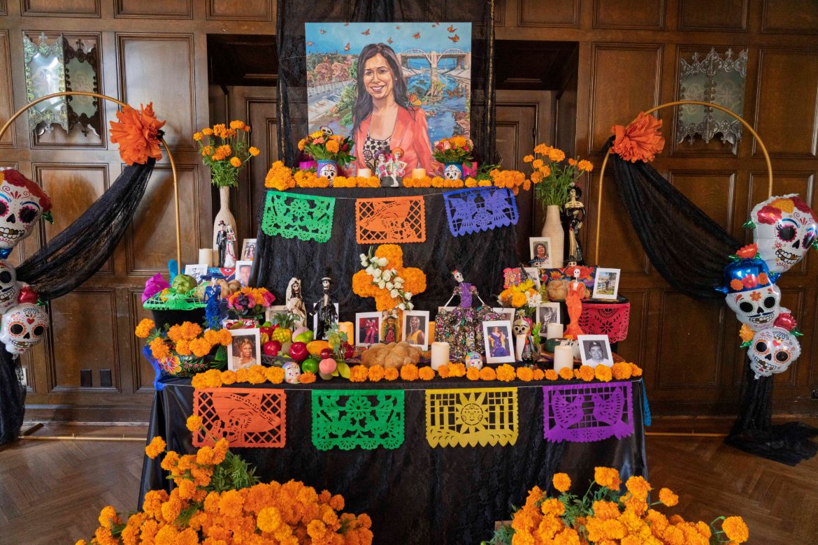A portrait of Cindy Montañez, standing in front of a river, sits atop a tiered ofrenda adorned with orange carnations and painted sugar skulls celebrating Día de los Muertos.