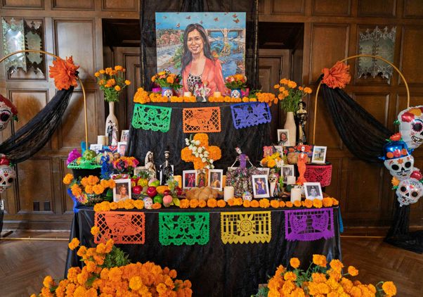 A portrait of Cindy Montañez, standing in front of a river, sits atop a tiered ofrenda adorned with orange carnations and painted sugar skulls celebrating Día de los Muertos.