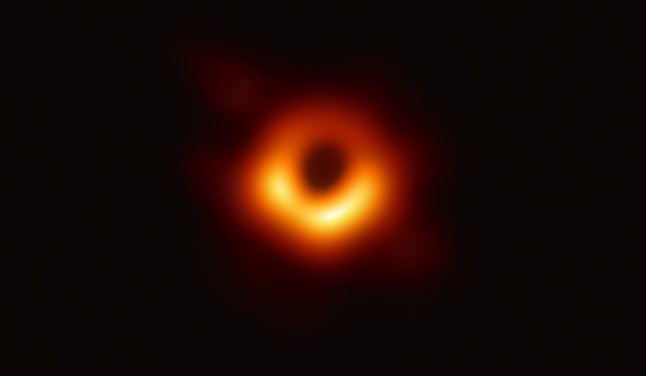 The first picture of a black hole, showing a black spot surrounded by a yellow-orange rim.