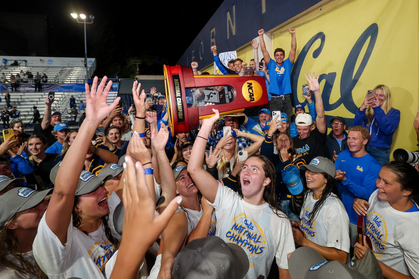 Members of UCLA's women's water polo team hold up NCAA championship trophy at poolside.