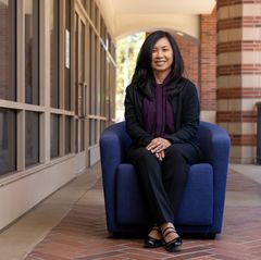 Gerrie Zvara sits smiling in a blue armchair along a brick colonnade corridor with large windows and red brick columns.
