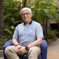 Kaya Mentesoglu wearing glasses and a light blue polo shirt sits smiling in a blue armchair surrounded by lush indoor greenery.