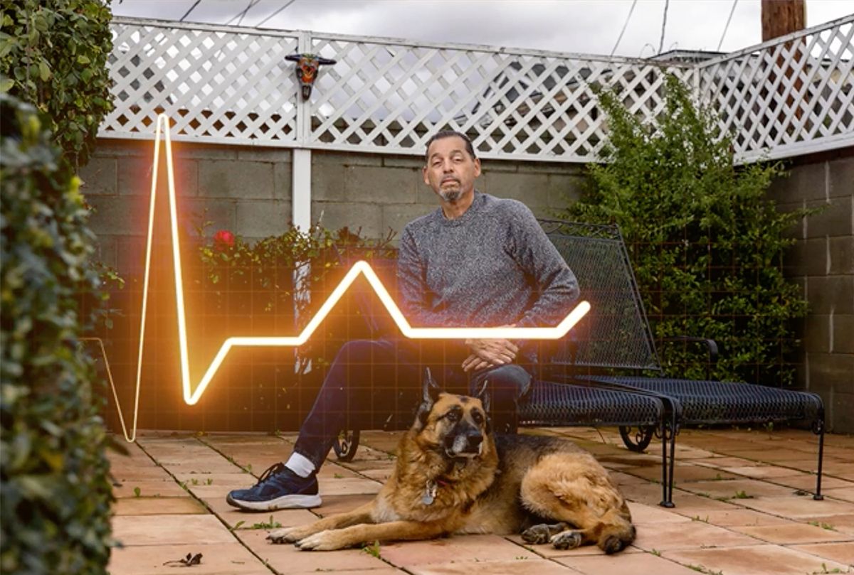 A man sits on a patio outdoors next to a large German Shepherd dog, with a glowing neon heartbeat line artwork behind him. The scene is surrounded by greenery and a white lattice fence.