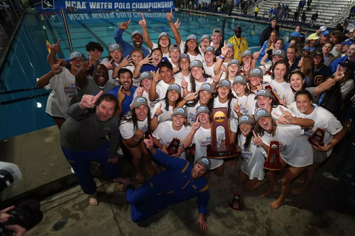 Athletes and coaches from UCLA’s women’s water polo team stand near pool with trophy and arms raised