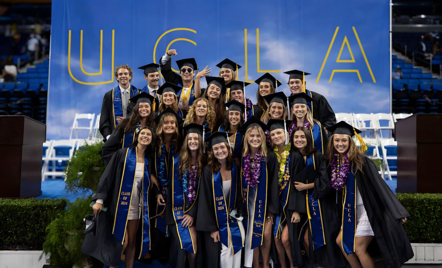 About two dozen graduates pose in front of UCLA sign in Pauley Pavilion