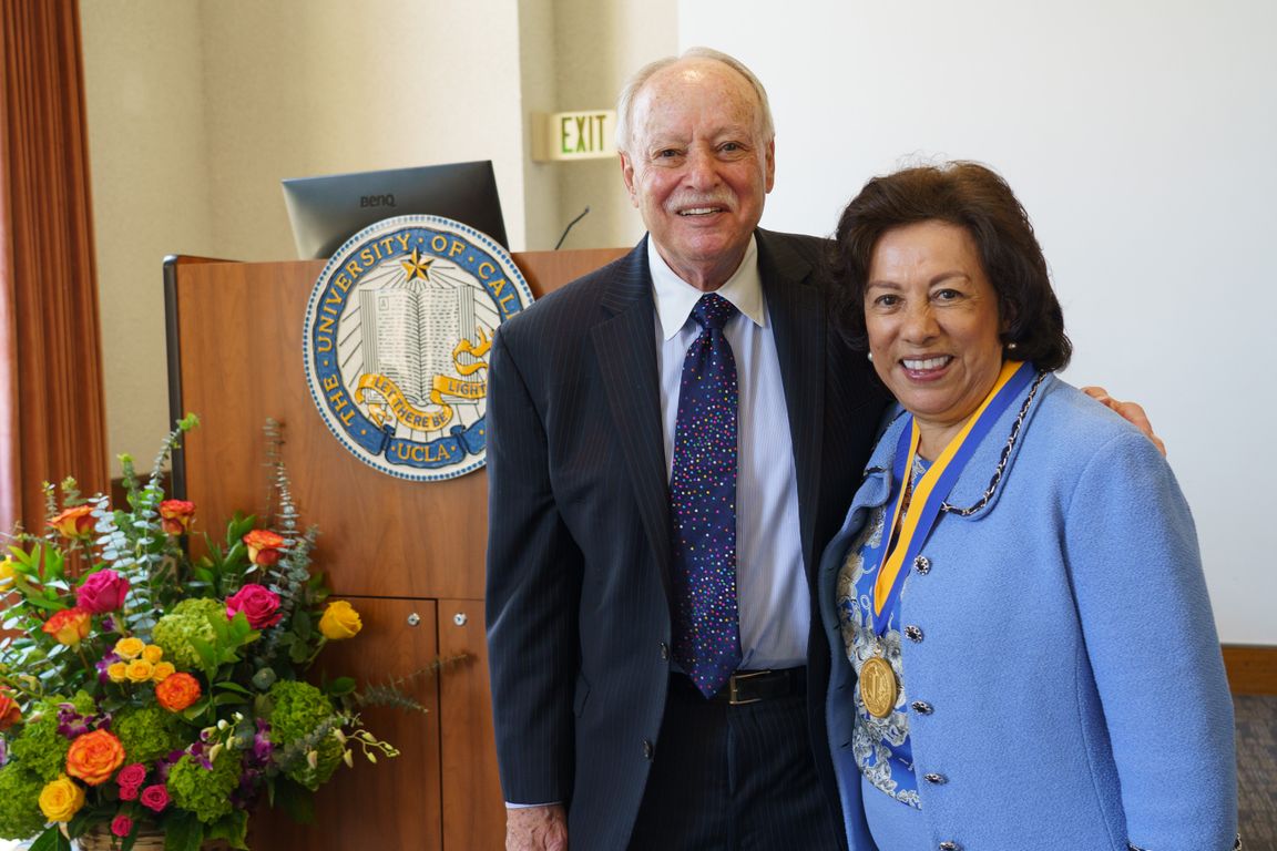 Antonia Hernández, right, and husband, Michael Stern, stand to right of podium; podium has UCLA seal and flower bouquet