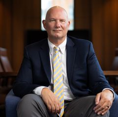 Joe Way in a navy blazer and striped tie sits smiling in a wood-paneled room with rows of chairs visible in the background.