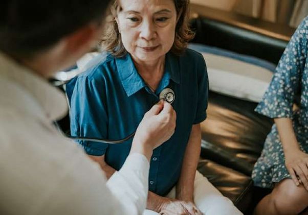 Seated older Asian woman in blue blouse and doctor checking her heart with stethoscope