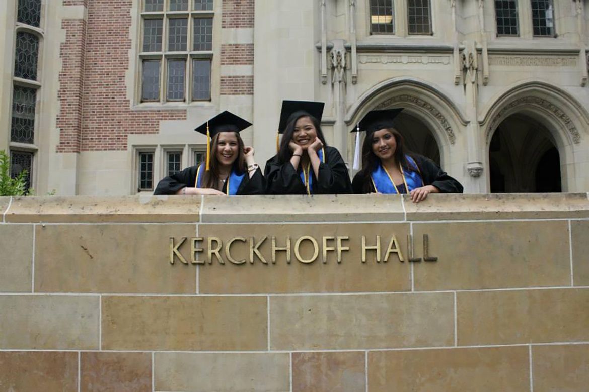 Three graduates in caps and gowns stand behind Kerckhoff Hall sign