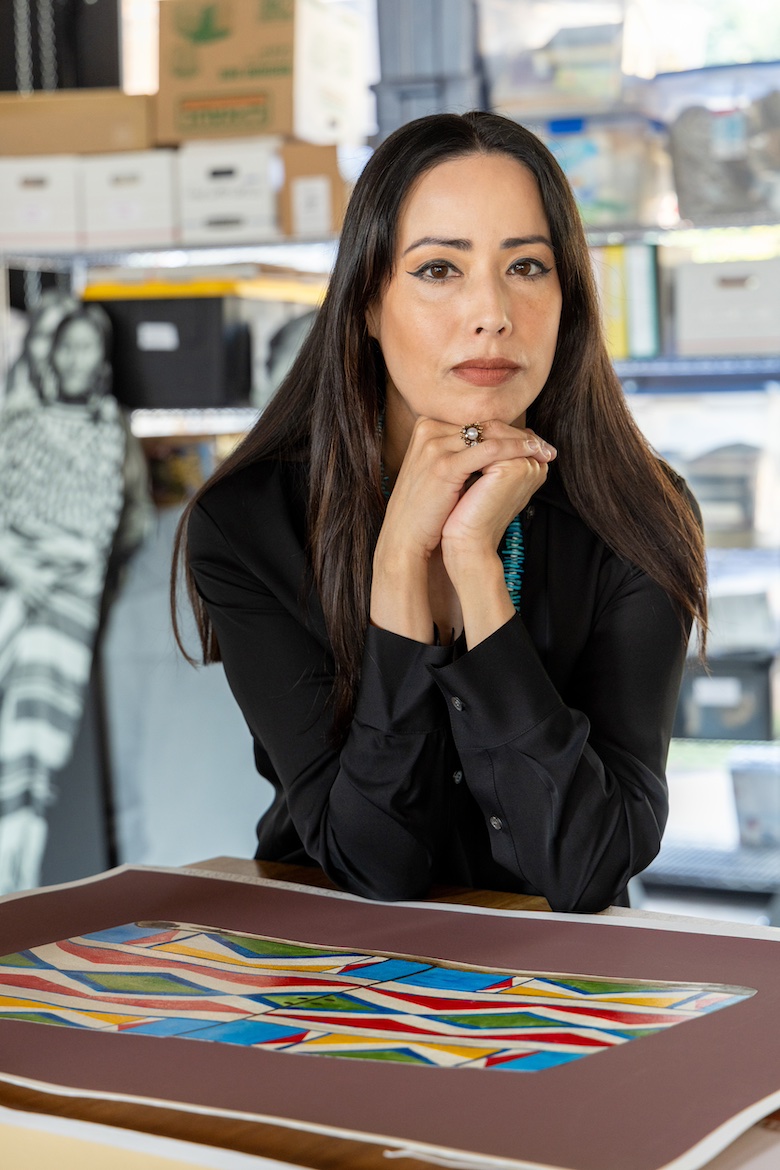 Artist Wendy Red Star with hands under chin sitting in front of a piece of art.