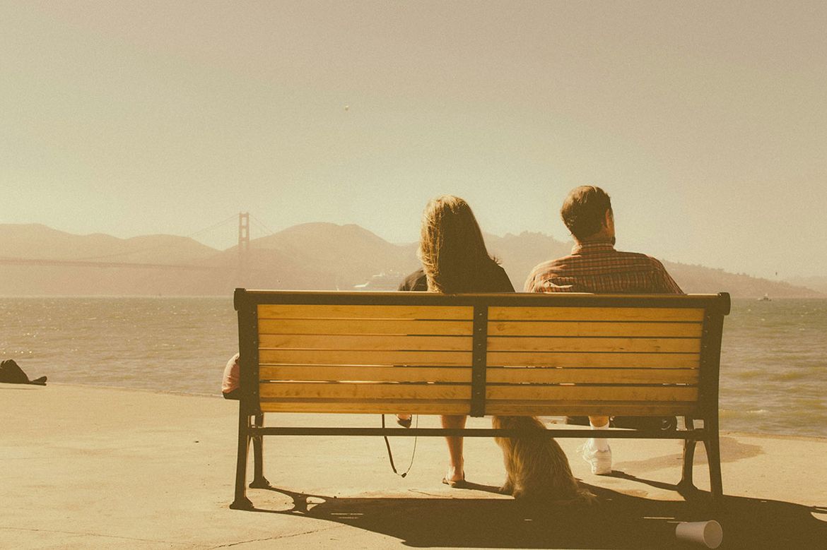 Couple sitting next to each other on a bench, with a dog between them, pictured from behind