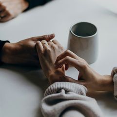 Two people hold hands across a white table, with a gray mug visible nearby, suggesting a moment of comfort or support.
