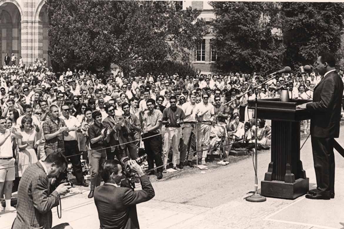 A black-and-white photograph of Martin Luther King Jr. speaking at a podium to a large crowd of students gathered outdoors at UCLA in 1965, about a month after the Selma to Montgomery march.
