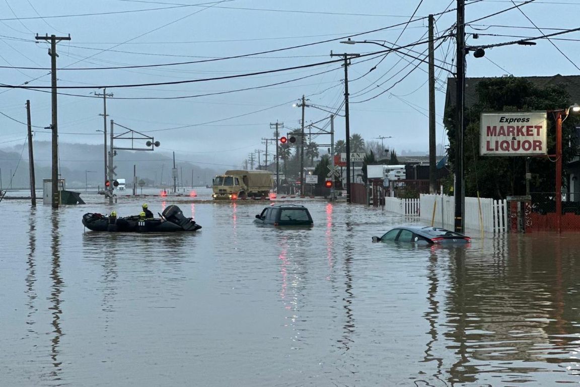 Autos sumergidos flotan en primer plano mientras un vehículo de la Guardia Nacional se acerca a una vía férrea inundada en el condado de Monterey California