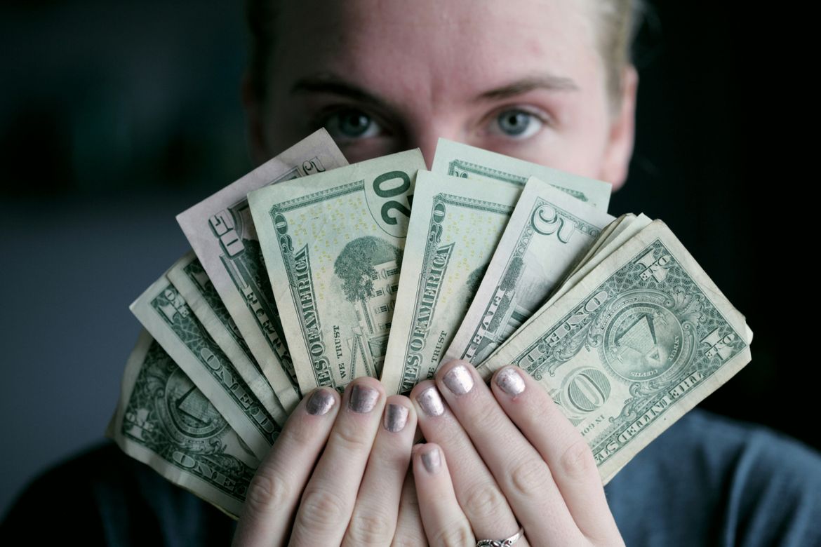A close-up image of a blue-eyed blonde-haired woman holding a fan of U.S. dollars