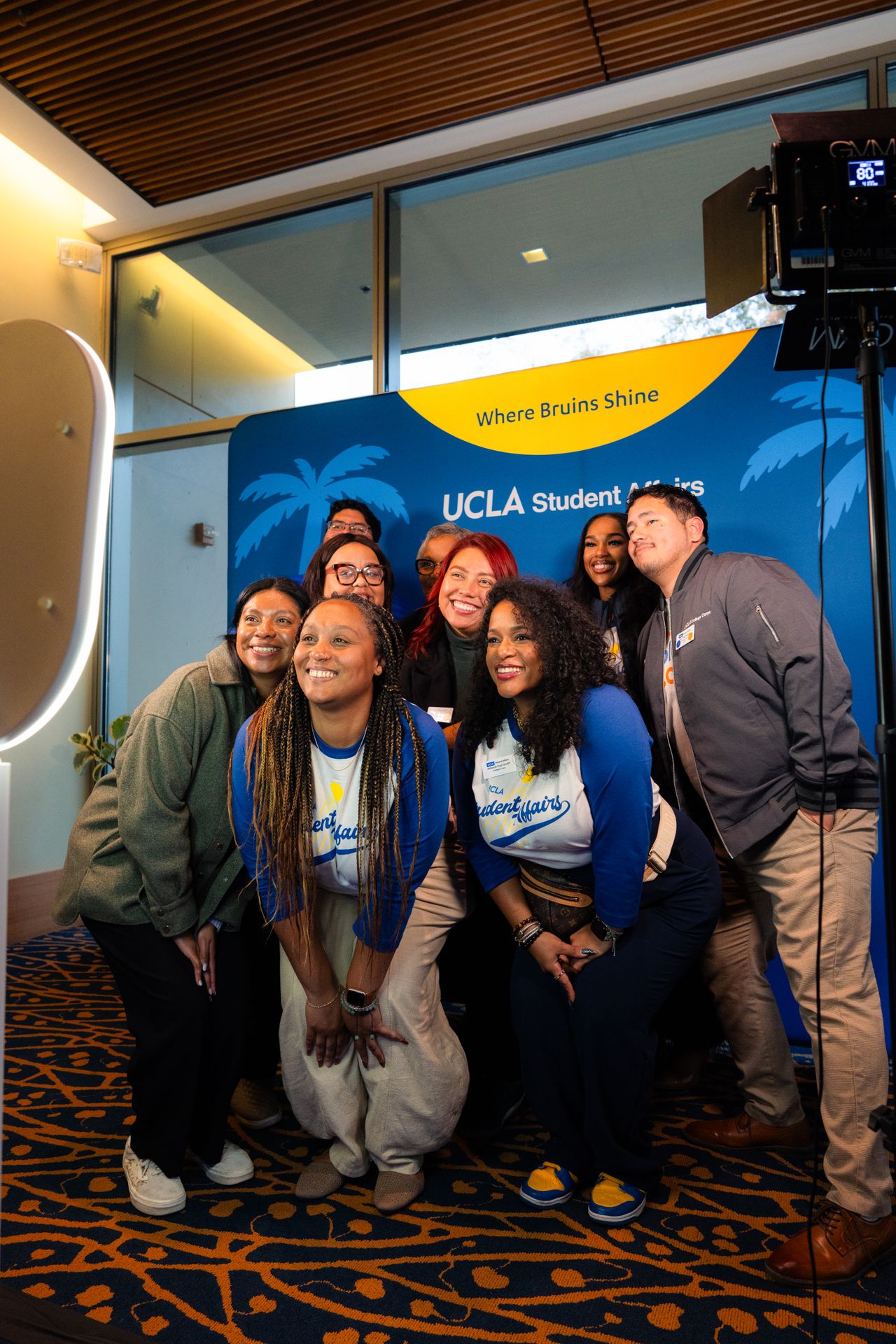 A group of nine people pose for a shot in front of a blue-and-gold sign that reads “Where Bruins Shine” and “UCLA Student Affairs”